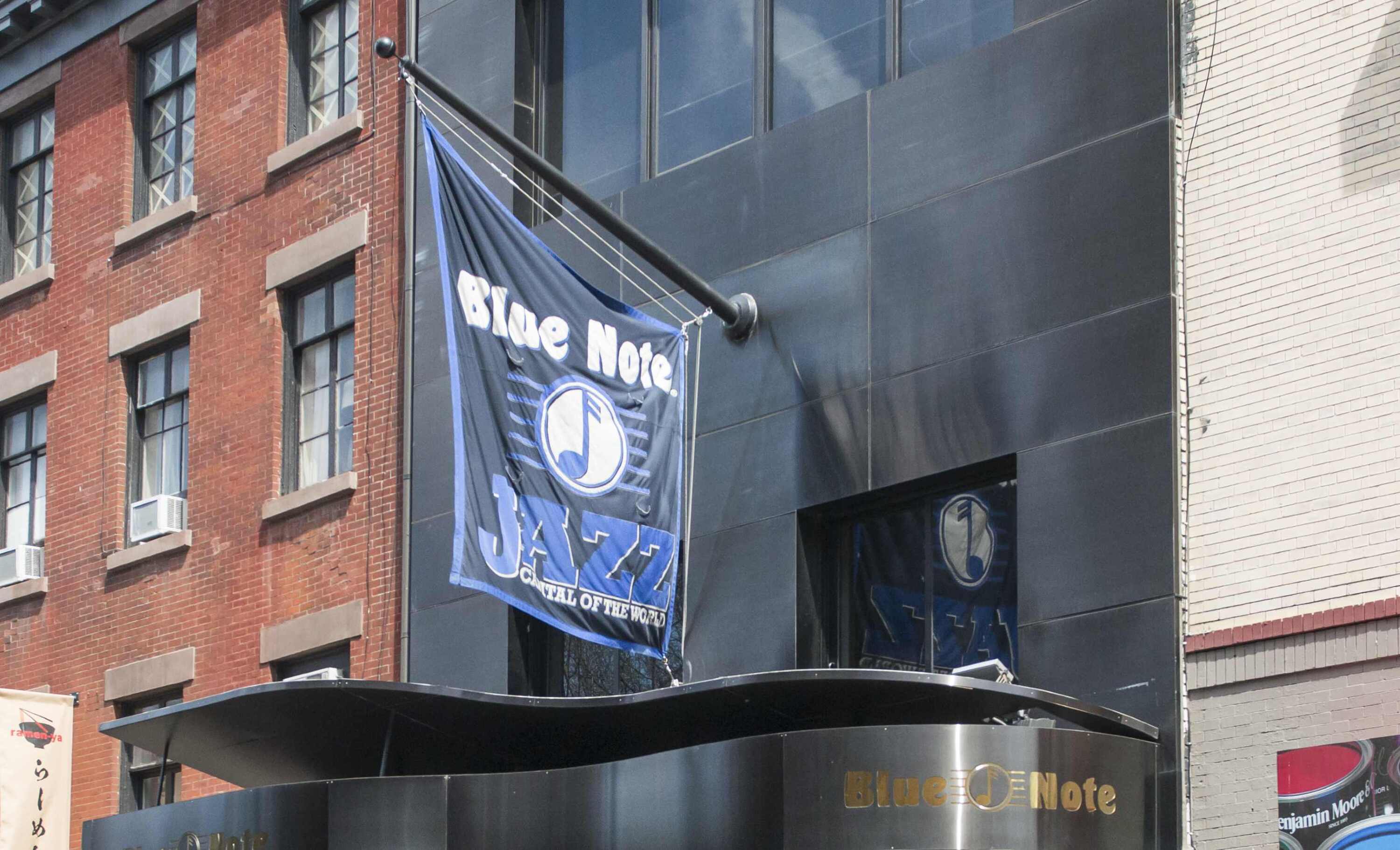 A Blue Note Jazz Club flag hangs outside a building with a modern black facade and adjacent brick structures, reflecting the club’s legal responsibility to maintain workplace safety for staff and guests.
