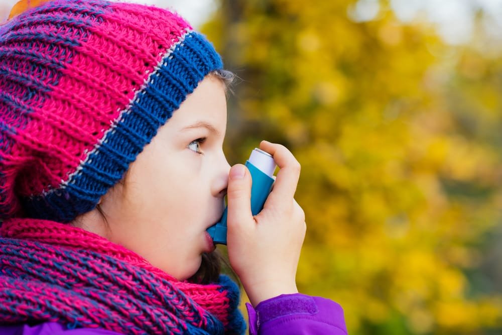 A little girl wearing a hat and scarf is using an asthma inhaler.