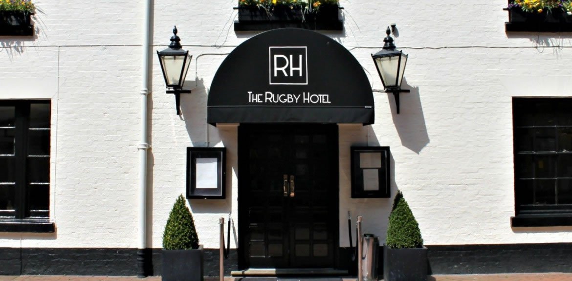 The entrance of The Rugby Hotel welcomes guests with its black doors, under a canopy boldly displaying "RH The Rugby Hotel." Two lanterns and symmetrically placed potted plants add charm, while inside, FREC3-trained staff ensure top-notch first response and emergency care services.